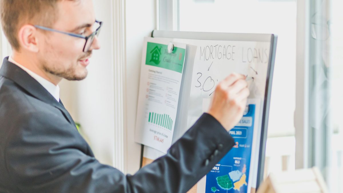 Businessman in suit writes on whiteboard displaying mortgage loan rates during a real estate meeting.