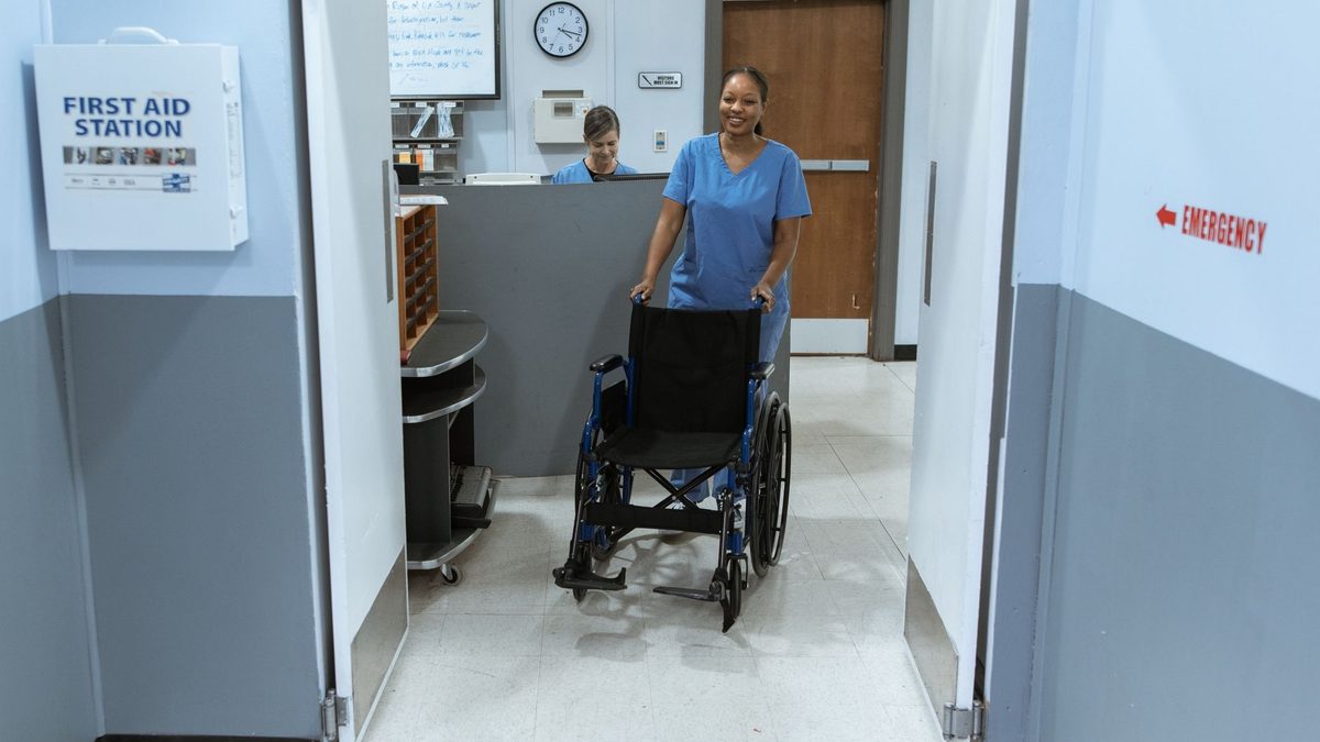 A nurse in scrubs pushes an empty wheelchair through a hospital hallway.